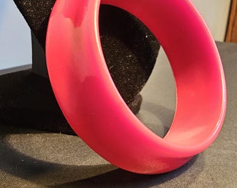 Può includere: Un braccialetto rigido in plastica rosa acceso. Il braccialetto è spesso e ha una finitura liscia e lucida.