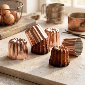 May include: A collection of copper canelé molds and baked canelés on a light-colored surface. The molds are empty and full, showcasing the fluted design. Other baking tools and ingredients are in the background, including eggs and a wire basket.