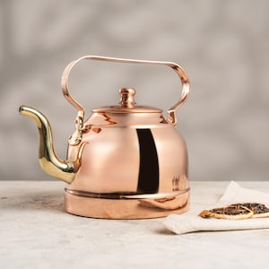 May include: A copper tea kettle with a gold-coloured spout and handle. The kettle has a rounded body and a lid with a small knob. The handle arches over the top. The kettle sits on a light-coloured surface.