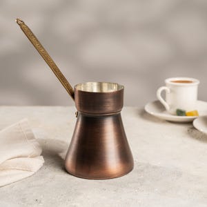 Puede incluir: Una cafetera turca de color cobre con un mango largo y adornado. La cafetera tiene una base ancha que se estrecha hacia un cuello más estrecho. Una taza y un platillo blancos están en el fondo.