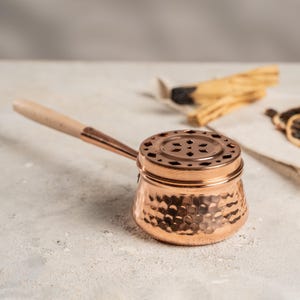 May include: A copper incense burner with a hammered finish and a wooden handle. The burner features a perforated lid and a small spout. The handle is made of light-coloured wood. The burner is designed for burning incense.