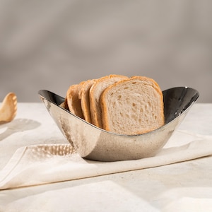 Könnte beinhalten: Ein silberfarbener, gehammerter Brotkorb aus Metall, gefüllt mit mehreren Scheiben Toastbrot. Der Korb hat eine einzigartige, gebogene Form und liegt auf einem weißen Tuch. Das Brot ist hellbraun mit goldener Kruste.
