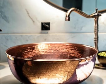Könnte beinhalten: Ein gehämmertes Kupferwaschbecken mit polierter Oberfläche. Das Waschbecken ist rund mit einem breiten Rand und steht auf einer weißen Arbeitsplatte. Ein Wasserhahn aus Kupfer und ein runder Spiegel befinden sich im Hintergrund.