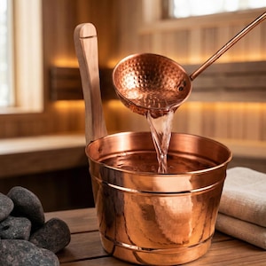 Könnte beinhalten: Ein Saunaeimer und Schöpflöffel-Set aus Kupfer. Der Eimer hat eine gehämmerten Oberfläche und einen Holzgriff. Wasser wird aus dem Schöpflöffel in den Eimer gegossen. Der Hintergrund zeigt einen hölzernen Saunainnenraum.