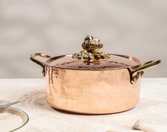 May include: Copper-colored cooking pot with a hammered texture and brass handles. The lid features a decorative frog-shaped handle. The pot is designed for kitchen use and adds a touch of elegance to any culinary space.