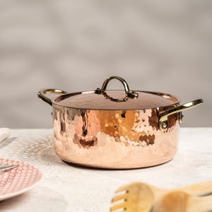 Può includere: Una pentola di rame con una texture martellata e un coperchio abbinato. La pentola ha due manici laterali e un manico superiore, tutti in color oro ottone. La pentola è su una superficie chiara.