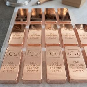 May include: Rows of copper bars, each stamped with "Cu", the atomic symbol for copper, and "ONE KILOGRAM 99.9 FINE COPPER". The bars are a warm, reddish-brown color, reflecting light. Additional tools and materials are visible in the background.