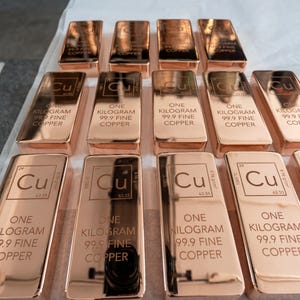 May include: Rows of polished copper bars, each stamped with "Cu" and "ONE KILOGRAM 99.9 FINE COPPER." The bars have a reflective, metallic finish, and are arranged neatly on a white surface. The image highlights the copper's reddish-brown color.