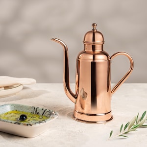 May include: A copper-colored oil dispenser with a long spout and a handle. The dispenser has a rounded body and a small lid. A small dish with olive oil and an olive is in the foreground, along with a sprig of olive leaves.