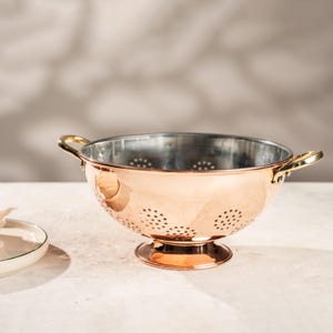 May include: A copper colander with a polished finish, featuring a footed base and two gold-toned handles. The interior is silver-coloured and perforated with small holes. A white plate with a green rim is in the background.