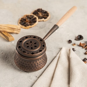May include: A copper incense burner with a wooden handle. The burner features a hammered texture and a decorative lid with a star-shaped design. Dried citrus slices and palo santo sticks are arranged nearby, suggesting aromatherapy and home fragrance.