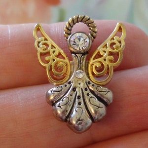 May include: A detailed angel pin with gold-colored filigree wings and a silver-colored body. The angel features a clear, round gemstone at the head and a smaller one on the body. The wings have an ornate, openwork design.