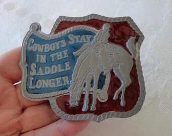 May include: A rectangular metal belt buckle with a cowboy riding a bucking horse. The buckle has a blue banner with the text "Cowboys Stay in the Saddle Longer." The border is red with a silver rope design.