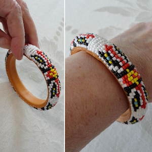 May include: A colourful beaded bracelet with a leather lining. The bracelet is decorated with white, red, black, and yellow beads. The bracelet is shown on a wrist and held by a hand.