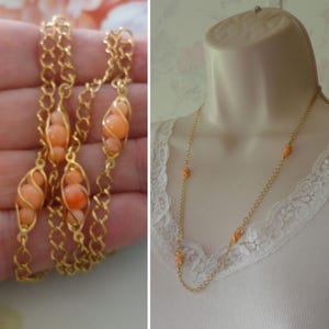 May include: Gold-toned necklace featuring peach-colored beads encased in delicate wirework. The necklace is displayed on a mannequin and held in a hand, showcasing its design and length. The chain has a delicate link pattern.