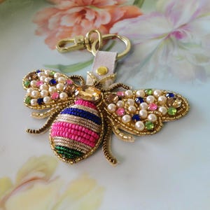 May include: A decorative bee keychain with gold-colored metal accents. The bee's body is pink and blue, with beaded wings adorned with pearls and colorful gems. The keychain includes a clasp and a leather strap.
