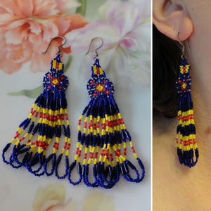 May include: Handmade beaded earrings in a vibrant color scheme. The earrings feature a blue, yellow, and red pattern. The top portion has a floral design, and the bottom has long beaded strands. The earrings are displayed on a white surface.
