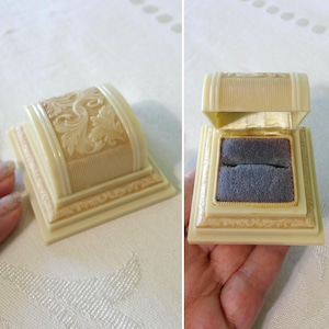May include: A cream-colored ring box with a hinged lid featuring an embossed floral scroll pattern. The exterior has a rectangular base with tiered edges. The interior is lined with a soft, dark blue velvet insert designed to hold a ring.