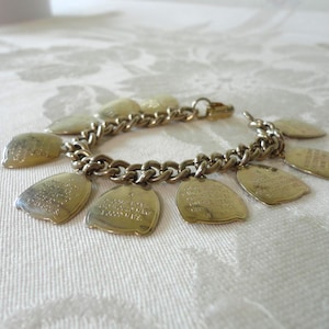 May include: A gold-tone metal charm bracelet featuring a series of shield-shaped pendants attached to a thick curb chain. Each pendant is engraved with text, and the bracelet is secured with a lobster claw clasp, showcasing a metallic finish.