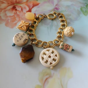 May include: A gold-tone double link charm bracelet featuring various pendants including a dark brown faceted stone, a round cream-colored piece with black calligraphy, a carved floral disc, a rectangular floral bead, and a smooth yellow bead.