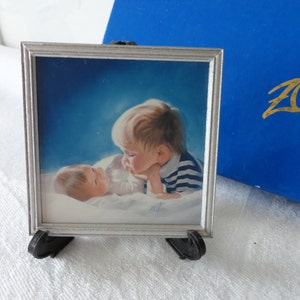 May include: A framed painting of two babies, one in a blue and white striped shirt, the other in white. The painting is in a silver frame and sits on a black easel. A blue box is in the background.