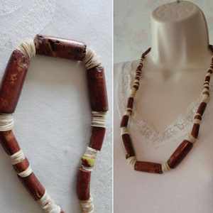 May include: A handmade necklace featuring alternating dark red cylindrical beads and small white shell-like beads. The necklace is displayed on a white surface and a mannequin, showcasing its design and length.
