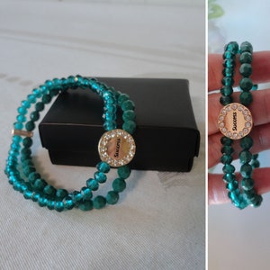 May include: A teal beaded bracelet with a gold-colored circular charm. The charm is encrusted with small clear crystals and features the word "Success" in black lettering. The bracelet is displayed on a white surface next to a black box.