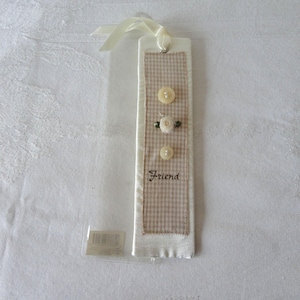 May include: A handmade bookmark with a beige and white gingham pattern, adorned with three buttons and a small fabric flower. The word "Friend" is embroidered on the bookmark. A cream-colored ribbon is attached at the top.
