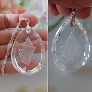 May include: Clear teardrop-shaped pendant necklace with a silver chain. The pendant features a detailed, etched seashell design. The pendant is held up by a silver bail. The necklace is displayed against a soft background.