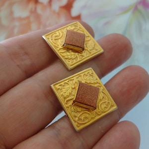 May include: A pair of gold-toned cufflinks with a square design. Each cufflink features a raised, square, copper-colored center stone set within a gold filigree frame. The frame has an ornate, floral pattern.