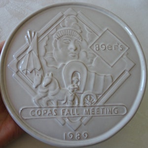 May include: Off-white ceramic plate with embossed relief design. Features a Native American profile, teepee, covered wagon, horse, and the text "89er's" and "COPAS FALL MEETING 1989".