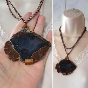 May include: A handcrafted necklace featuring a large, irregularly shaped pendant. The pendant showcases a dark blue and brown agate stone, accented with copper-colored metal. The necklace has a double strand chain, creating a unique and eye-catching accessory.