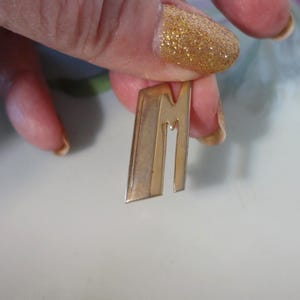 May include: A gold-colored metal pin in the shape of the letter "M". The pin has a rectangular shape with a vertical line and a stylized "M" cut out. The pin is held between fingers.