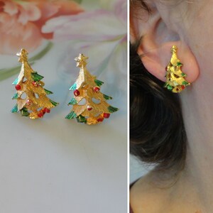 May include: Gold-toned Christmas tree earrings with red and green accents. The earrings are shaped like stylized Christmas trees, complete with a star on top. The earrings are shown on a white surface and on an ear.