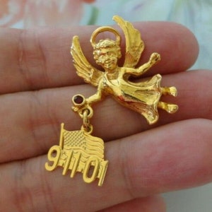 Peut inclure: Une broche dorée en forme d'ange aux bras et aux ailes déployés. L'ange tient un petit charme avec un drapeau américain flottant et la date "9-11-01". La broche a une auréole et une petite gemme rouge.