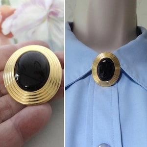 May include: A circular brooch featuring a polished black oval cabochon stone set in the center of a gold-toned metal frame. The frame is designed with concentric circular ridges that radiate outward from the central black stone.