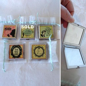 May include: A collection of square, silver-framed compact mirrors with various designs and quotes. Some mirrors are in sheer pouches with blue ribbons. One mirror is open, revealing the mirrored surfaces.