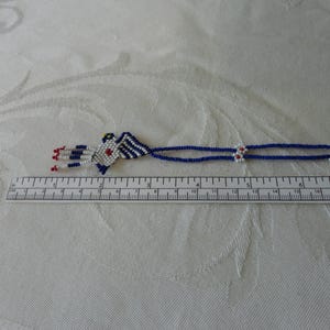 Pu&ograve; includere: Una collana di perline con un design geometrico. La collana presenta un ciondolo centrale con perline bianche, blu e rosse. La collana &egrave; lunga circa 33 cm. Lo sfondo &egrave; un tessuto bianco fantasia.