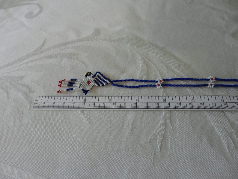Pu&ograve; includere: Una collana di perline con un design geometrico. La collana presenta un ciondolo centrale con perline bianche, blu e rosse. La collana &egrave; lunga circa 33 cm. Lo sfondo &egrave; un tessuto bianco fantasia.