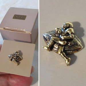 May include: A vintage gold-tone metal Avon football player pin. The pin shows a football player running with the ball. The pin is on a cream-coloured square, with a matching box with the Avon logo.