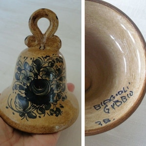 May include: A ceramic bell with a loop handle, painted with a floral design in shades of blue and black on a tan background. The bell's interior is tan with brown trim and has handwritten text.