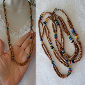 May include: A long, layered necklace featuring small, light brown wooden beads interspersed with colorful glass beads. The glass beads are in shades of red, yellow, blue, green, and black. The necklace is displayed on a white surface.