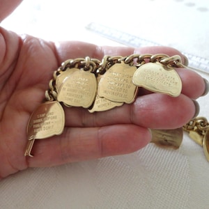 May include: A gold-toned charm bracelet with multiple rectangular and leaf-shaped charms. Each charm has engraved text. The bracelet is held in a hand. The charms have phrases like "Stop and think before you drink - then don't."