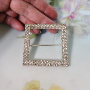 May include: A square-shaped brooch featuring a silver-toned metal frame completely encrusted with small, clear, sparkling rhinestones. The center of the square is open, and a thin metal pin mechanism is attached horizontally across the back.
