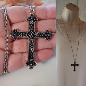 May include: A silver-toned cross pendant necklace with an ornate, detailed design. The cross features a black background with floral and dot patterns. The necklace is displayed on a hand and a mannequin.