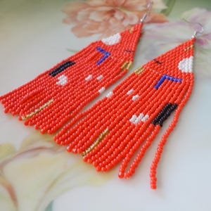 May include: A pair of long, triangular beaded earrings featuring a vibrant orange base color. The design incorporates small, scattered geometric accents in white, black, blue, and metallic gold beads, finished with silver-toned metal earring hooks.