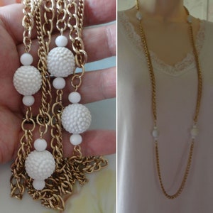 May include: Gold-tone chain necklace with white textured and smooth beads. The necklace is shown in two views, one held in a hand and the other displayed on a mannequin. The necklace has a long, layered design.