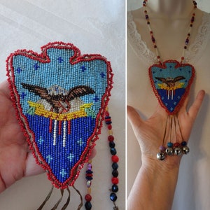 May include: A beaded pendant necklace with a heart-shaped design. The pendant features a blue background with an eagle, red trim, and dangling beads. The necklace has a beaded chain and small silver bells.
