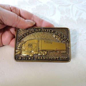 May include: A rectangular brass belt buckle featuring a raised relief of a semi-truck. The text DRIVERS SERVICE AWARD is arched across the top, and J.B. HUNT TRANSPORT, INC. is embossed along the bottom. The background has a textured finish.