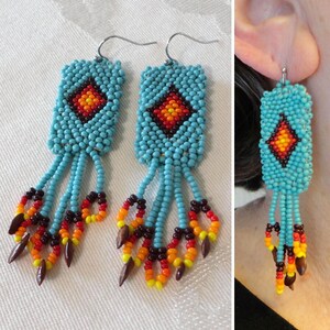May include: Turquoise beaded earrings with a diamond pattern in red, orange, and yellow. The earrings have dangling beaded strands in turquoise, red, orange, and yellow, with brown teardrop-shaped accents. The earrings are handmade.